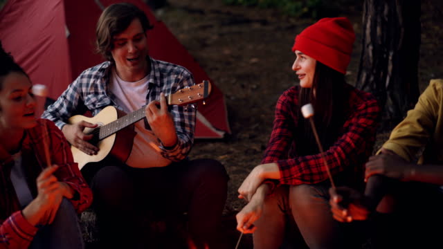 Friends-at-campsite-are-singing-songs,-laughing-and-cooking-food-on-fire-while-happy-smiling-guy-is-playing-the-guitar.-Camping,-music-and-friendship-concept.