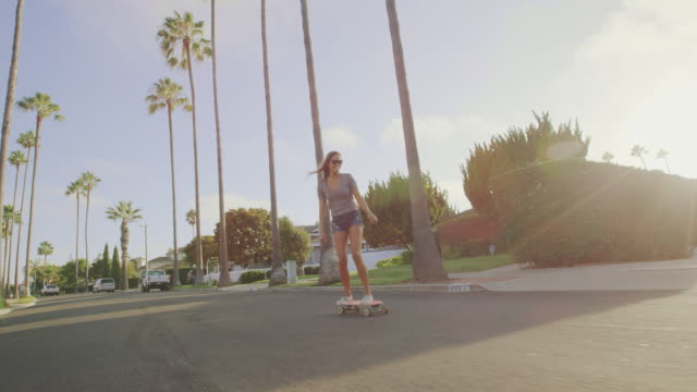 Attractive-adventurous-girl-skateboarding-down-palm-tree-lined-street-at-sunset