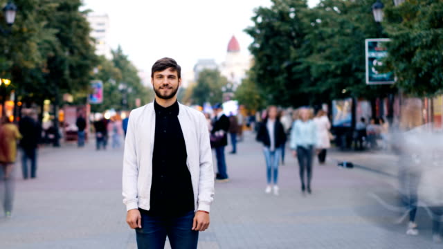 Time-lapse-of-confident-man-in-jeans-and-white-jacket-standing-alone-in-street-in-city-and-looking-at-camera-while-people-are-rushing-by-on-autumn-day.