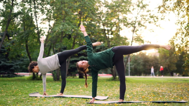Beautiful-young-ladies-are-standing-in-Half-Moon-pose-Ardha-Chandrasana-during-pair-yoga-class-in-park.-Healthy-lifestyle,-teaching-and-learning-and-sports-concept.