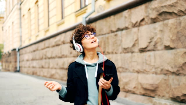 Gut-aussehende-Brünette-in-stilvollen-Gläsern-ist-Radio-im-Kopfhörer-hören-und-mit-Smartphone-beim-Spaziergang-in-Modent-Stadt-im-Herbst.-Menschen-und-urban-Life-Konzept.