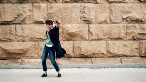 Excited-girl-student-is-dancing-and-singing-enjoying-music-in-headphones-standing-outdoors-with-stone-wall-in-background.-Young-woman-is-holding-smartphone.