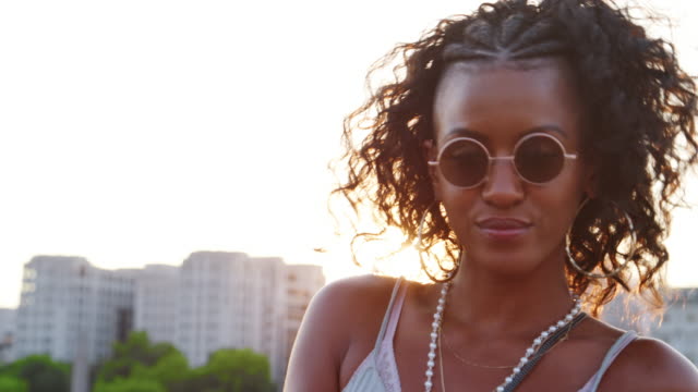 Junge-schwarze-Frau-mit-Sonnenbrille-auf-Kamera-und-lächelnd-auf-einer-Londoner-Straße-bei-Sonnenuntergang-hautnah