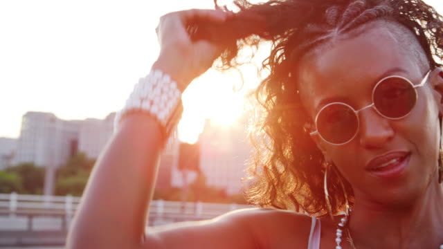 Close-up-of-young-black-woman-wearing-sunglasses-plying-with-her-hair,-looking-to-camera-on-a-London-street-at-the-golden-hour