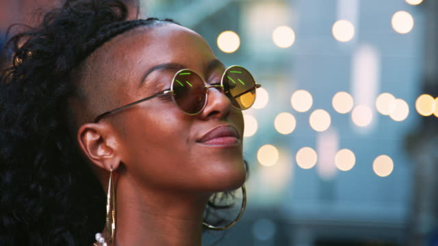 Young-black-woman-looking-up,-turns-to-camera-and-takes-off-sunglasses,-head-shot,-bokeh-lights-in-background