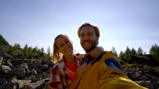 Hikers,-couple--taking-selfie-portrait-on-mountain-trail.-sun-shining-over-the-mountains.-Beautiful-sunbeam-effect-making-an-idyllic-landscape.-Young-couple-hiking-take-video-selfie-in-Switzerland