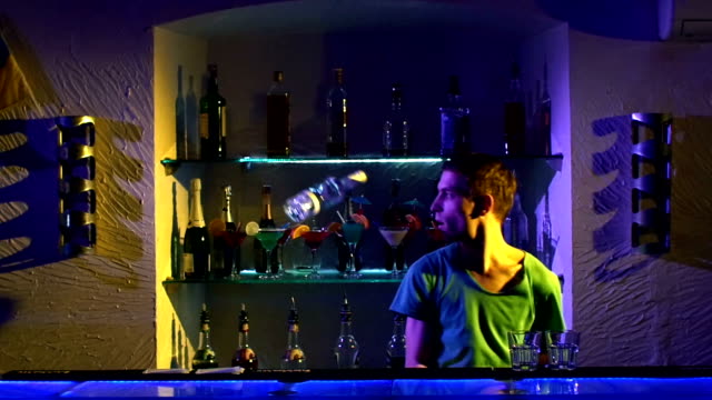 Young-bartender-making-tricks-with-two-glasses-and-bottle--standing-behind-the-bar,-catching,-throwing-up-slow-motion