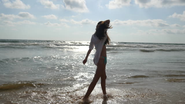 Mujer-feliz-en-bikini-y-camiseta-caminando-en-el-agua-de-mar-en-la-playa.-Joven-hermosa-chica-disfrutando-de-la-vida-y-la-diversión-a-la-orilla-del-mar.-Vacaciones-de-verano-o-vacaciones
