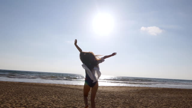 Young-woman--running-on-beach-to-the-ocean-at-sunset-and-raised-hands.-Beautiful-young-girl-going-on-sandy-shore-to-the-sea-and-enjoying-freedom-during-vacation.-Relax-on-summer-holiday.-Rear-back-view