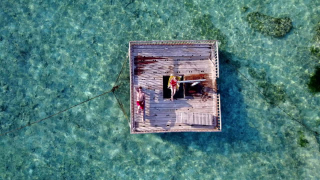 v03858-Aerial-flying-drone-view-of-Maldives-white-sandy-beach-2-people-young-couple-man-woman-relaxing-on-sunny-tropical-paradise-island-with-aqua-blue-sky-sea-water-ocean-4k-floating-pontoon-jetty-sunbathing-together