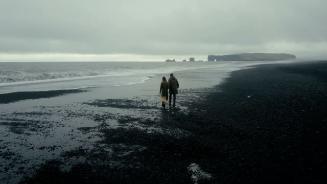 Aerial-view-of-young-stylish-couple-walking-on-the-black-volcanic-beach-in-Iceland.-Man-and-woman-running-from-the-wave