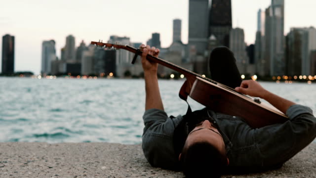 Elegante-joven-acostado-en-la-orilla-del-lago-Michigan-en-Chicago,-América-y-tocando-la-guitarra-acústica