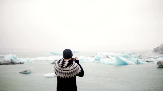 Back-view-of-young-traveling-man-standing-in-Jokulsalon-ice-lagoon-in-Iceland-and-take-photos-og-glaciers-on-smartphone