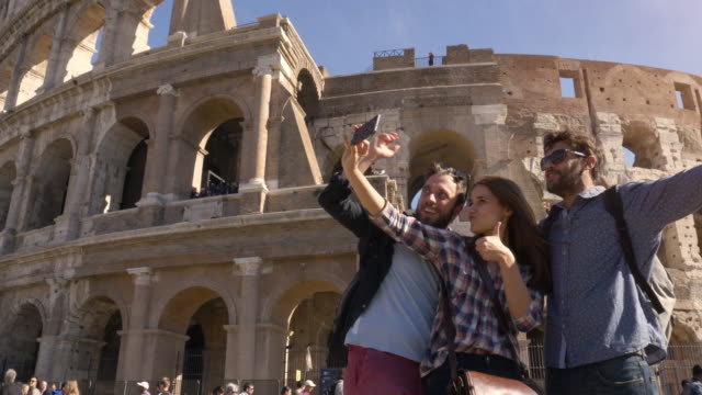 Three-young-friends-tourists-standing-in-front-of-colosseum-in-rome-taking-selfies-with-smartphone-with-backpacks-sunglasses-happy-beautiful-girl-long-hair-slow-motion
