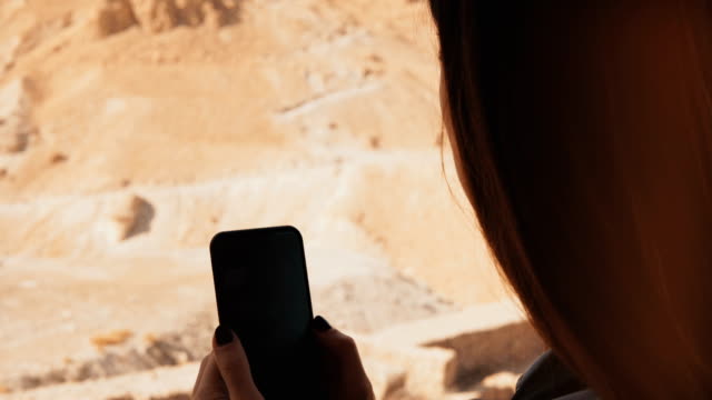 Girl-using-smartphone-in-aerial-railway-cabin.-Woman-takes-phone-photos-in-sunny-desert-ropeway-car.-Masada,-Israel.-4K