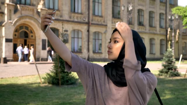 Young-beautiful-muslim-girl-in-hijab-is-making-selfie-on-her-smartphone-in-daytime-in-summer,-building-on-background,-religiuos-concept,-communication-concept