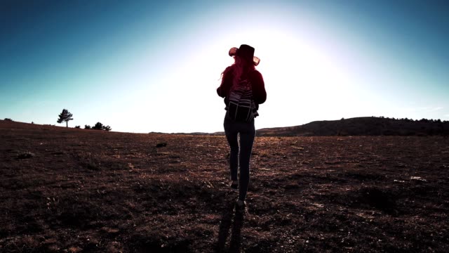 Silhouette-of-slender-sports-female-in-hat-with-backpack-walking-on-field-at-sunset