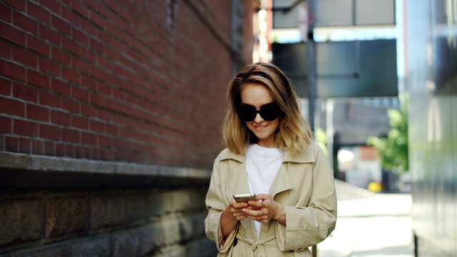 Hübsche-Blondes-Mädchen-nutzt-moderne-Smartphone-Bildschirm-Wandern-in-Stadt-und-Lächeln-auf-den-Lippen-zu-berühren.-Junge-Frau-trägt-modische-Kleidung-und-trendige-Sonnenbrillen.
