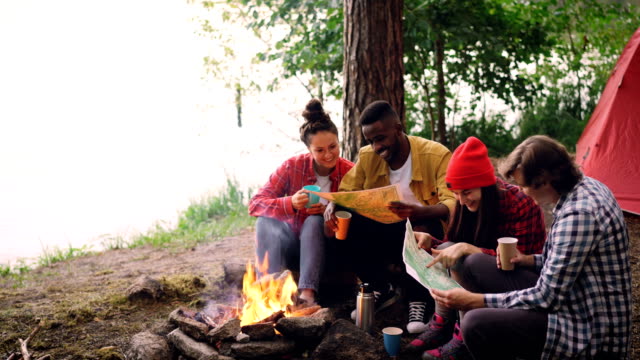 Cinemagraph-loop---group-of-young-men-and-women-are-looking-at-paper-maps-sitting-around-fire-in-forest-with-drinks.-Map-is-moving,-tent-and-trees-are-visible.