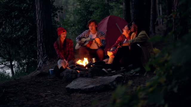 Cheerful-travelers-are-singing-songs-and-playing-the-guitar-sitting-around-fire-in-forest-in-the-evening-and-having-fun-with-beautiful-nature-around.-People-and-music-concept.
