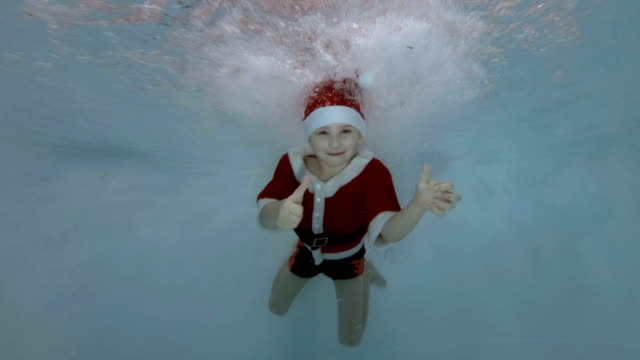 Un-niño-vestido-de-Santa-Claus-nada-bajo-el-agua-en-los-chorros-de-agua-en-la-piscina-con-su-sonrisa-abierta,-ojos,-mira-a-la-cámara-y-demuestra-sus-dedos-para-arriba.