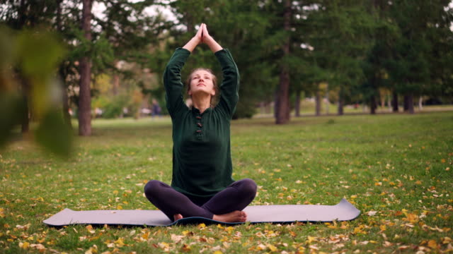 Good-looking-girl-is-sitting-in-lotus-position-on-yoga-mat-on-grass-in-city-park-holding-hands-in-namaste-then-in-mudra-on-knees-and-breathing.-Meditation-and-nature-concept.