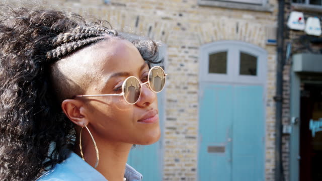 Fashionable-young-black-woman-wearing-blue-dress-and-sunglasses-looking-around-on-the-street,-side-view,-close-up