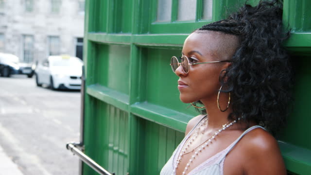 Trendige-junge-schwarze-Frau-tragen-Runde-Sonnenbrille-und-Leibchen-stützte-sich-auf-grüne-Wand-in-einer-Stadtstraße,-Seitenansicht,-Kopf-und-Schultern