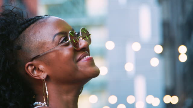 Fashionable-young-black-woman-in-sunglasses-looking-around,-head-shot,-bokeh-lights-in-background
