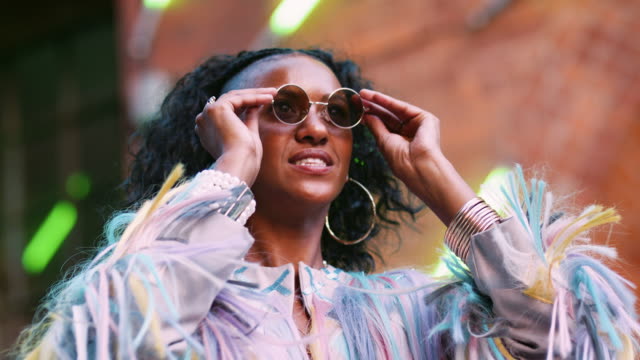Young-black-woman-wearing-multicoloured-fringed-jacket-putting-on-sunglasses,-low-angle,-head-and-shoulders