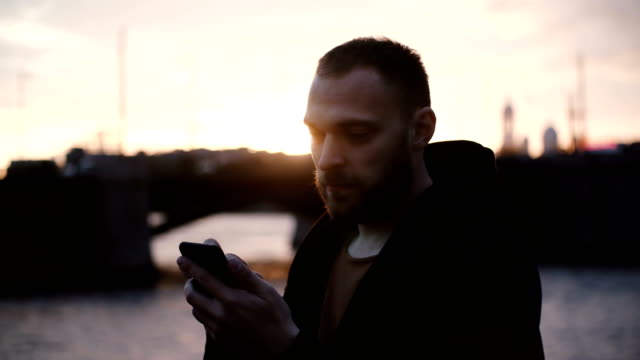 Portrait-of-young-bearded-man-spending-time-on-beautiful-embankment,-using-the-smartphone-in-evening-on-sunset