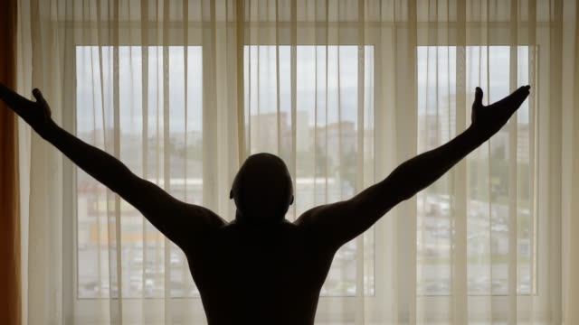 Adult-white-caucasian-sports-men-standing-in-front-of-window-with-raised-hands-at-sunrise.-Men-silhouette.-He-feels-free-and-happy.-Force-and-energy-overflows-his-body.