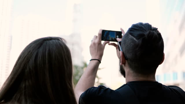 Young-beautiful-couple-walking-in-downtown-of-Chicago,-America-and-taking-photo-of-skyscraper-on-smartphone