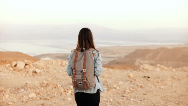 Woman-with-backpack-walks-in-cloudy-desert.-Young-thoughtful-Caucasian-girl-wanders-on-dry-sand,-smiling.-Israel-4K