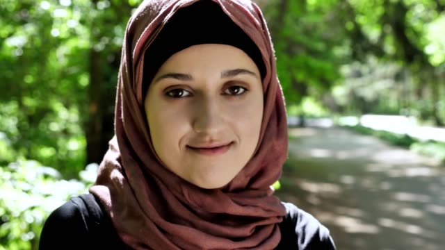 Porträt-von-einem-netten-jungen-Mädchen-in-einem-Hijab,-lächelt,-schaut-in-die-Kamera,-Park-im-Hintergrund.-50-fps