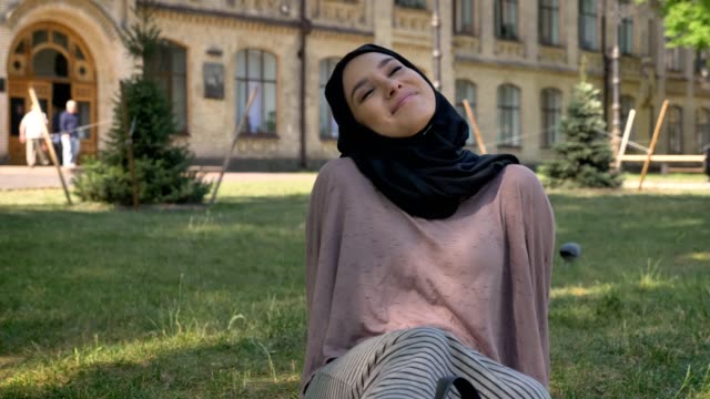 Junge-schöne-muslimische-Mädchen-Träumer-Hijab-sitzt-auf-Rasen,-Builging-auf-Hintergrund,-religiöse-Konzept,-relax-Konzept