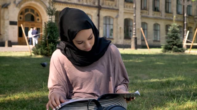 Niña-musulmana-en-hiyab-está-sentada-sobre-césped-y-leer-revista,-Hausmanie-en-el-fondo,-concepto-religioso,-relajarse-concepto