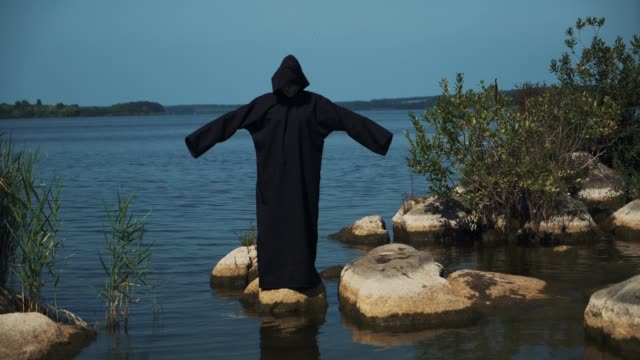 Woman-devil-in-black-cape-and-hood-on-stones-near-the-river.