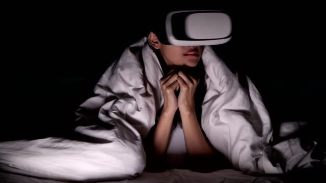 Woman-watching-VR-horror-movie-alone-in-bed.-Asian-woman-lying-in-bed-alone-wearing-VR-head-set,-very-surprise-and-shock-from-scary-movie.-High-tech-Halloween-home-alone-concept.