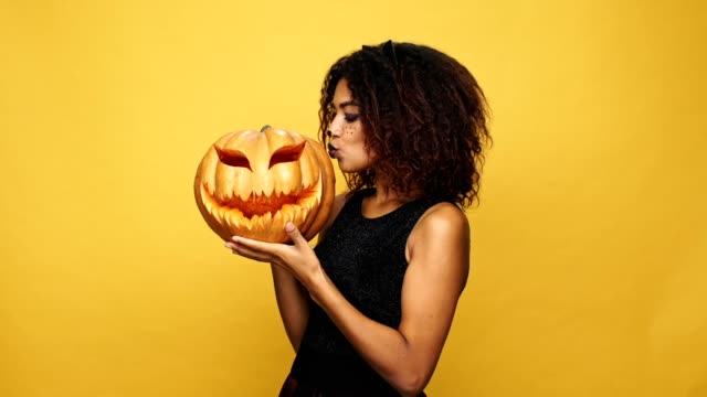 Funny-african-lady-in-halloween-costume-showing-and-playing-with-pumpkin-isolated-over-yellow-background