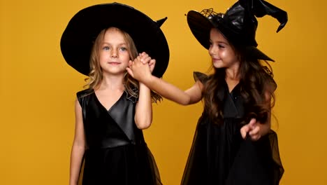 Kleine-hübsche-Freunde-Mädchen-in-schwarz-Hexen-Kleider-und-Hüte,-Hand-in-Hand-miteinander-und-lächelnd-über-orange-Hintergrund-dekoriert