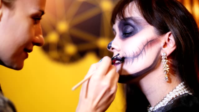Fiesta-de-Halloween,-Maquillaje-artista-dibuja-un-maquillaje-terrible-en-la-cara-de-una-mujer-morena-para-una-fiesta-de-Halloween.-en-el-fondo,-se-ve-el-paisaje-en-el-estilo-de-Halloween