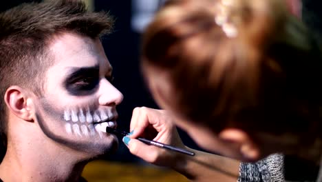 Halloween-partido,-primer-plano,-Maquillaje-artista-dibuja-un-maquillaje-terrible-en-la-cara-de-un-hombre-para-una-fiesta-de-Halloween.-en-el-fondo-se-ve-el-paisaje-en-el-estilo-de-Halloween