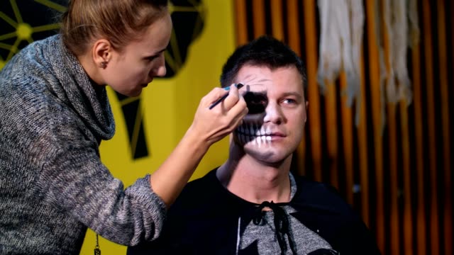 Halloween-party,-make-up-artist-draws-a-terrible-makeup-on-the-face-of-a-man-for-a-Halloween-party.-in-the-background,-the-scenery-in-the-style-of-Halloween-is-seen