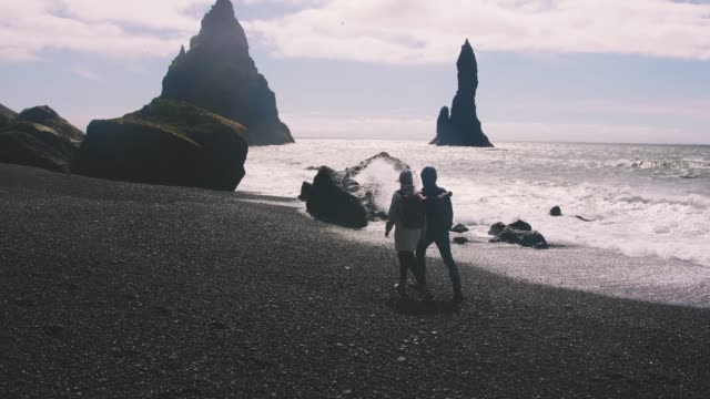 Young-couple-holding-hands-walking-down-black-sand-beach-in-Iceland,-slow-motion