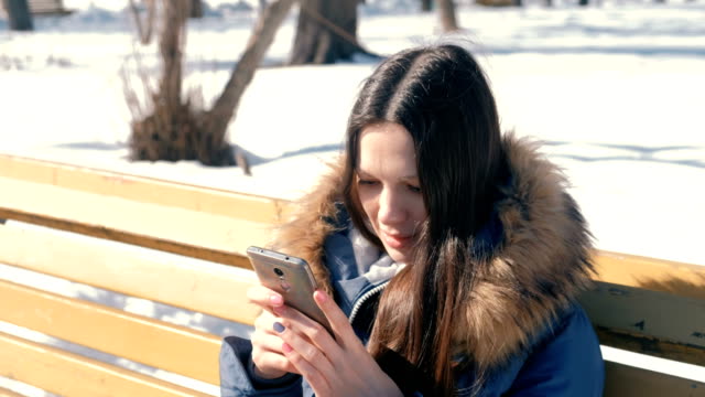 Happy-young-woman-brunette-types-a-message-on-her-phone-sitting-on-the-bench-in-winter-city-park-in-sunny-day.