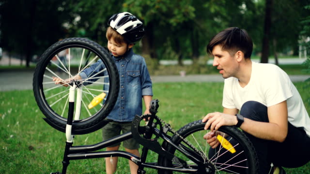 Neugieriges-Kind-tragen-Helm-dreht-Rad-und-Pedale,-während-sein-Vater-auf-Rasen-im-Park-am-Sommertag-im-Gespräch-mit-ihm-ist.-Familie,-Freizeit-und-aktiven-Lifestyle-Konzept.