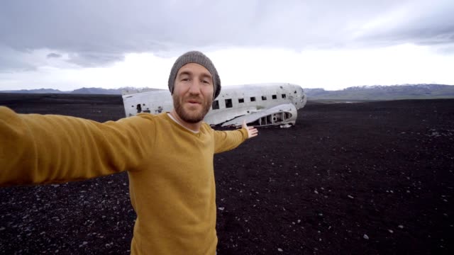 4k-Jüngling-durch-Flugzeug-Wrack-am-schwarzen-Sandstrand-unter-einen-Selfie-Porträt-berühmten-Platz-stehen-zu-Besuch-in-Island-und-ist-mit-dem-Wrack