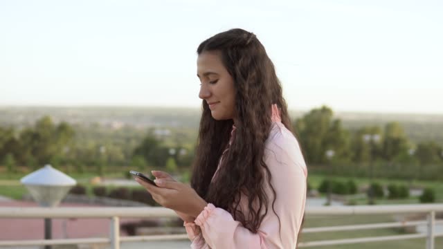 A-girl-with-long-hair-walks-in-the-park-near-the-Ferris-wheel-and-uses-a-smartphone.-4K