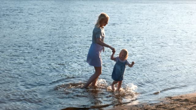 Mom-and-girl-walking-on-the-beach-of-the-river-at-sunset-and-swim-in-city.